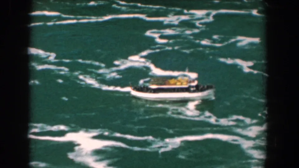 1950s 8mm frame of a small tour boat crossing turbulent green water with white foam patterns around it.