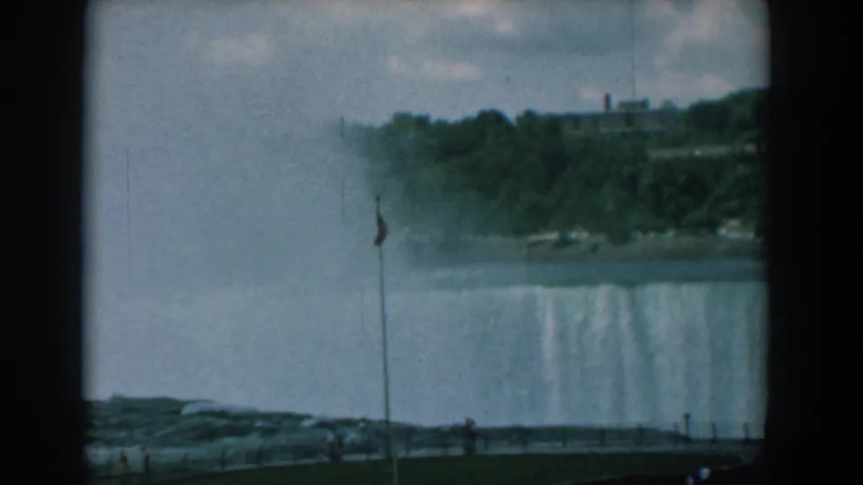 Wide 8mm view toward a large waterfall with mist, a foreground flagpole, tiny visitors, wooded cliffs, and a distant building.