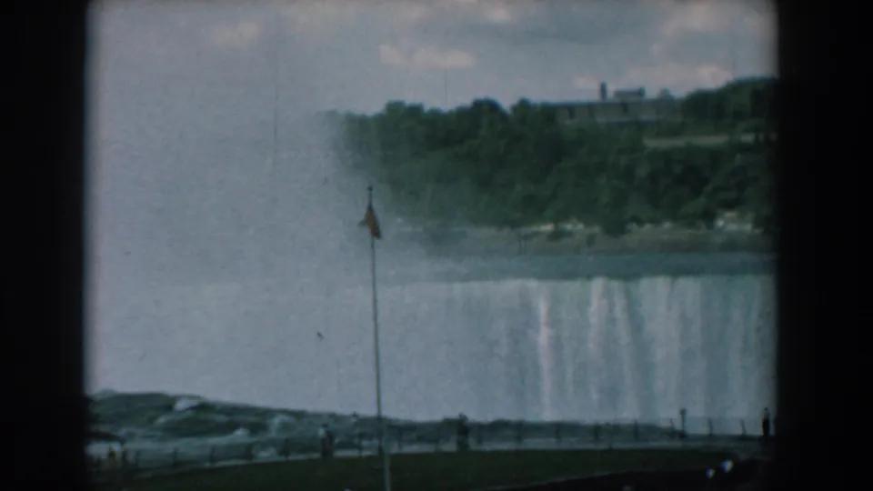 The falls remain in view from an overlook, with a flagpole, mist, wooded cliffline, and tiny visitors near the lower edge.