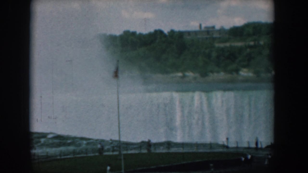 1958 8mm travel frame showing a broad waterfall, mist, an American flag, trees, and distant visitors on an overlook path.