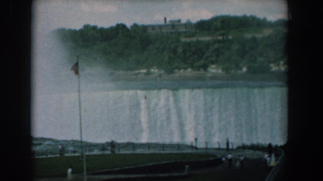 Waterfall and mist viewed past a flagpole, with tourists on a curved overlook path.