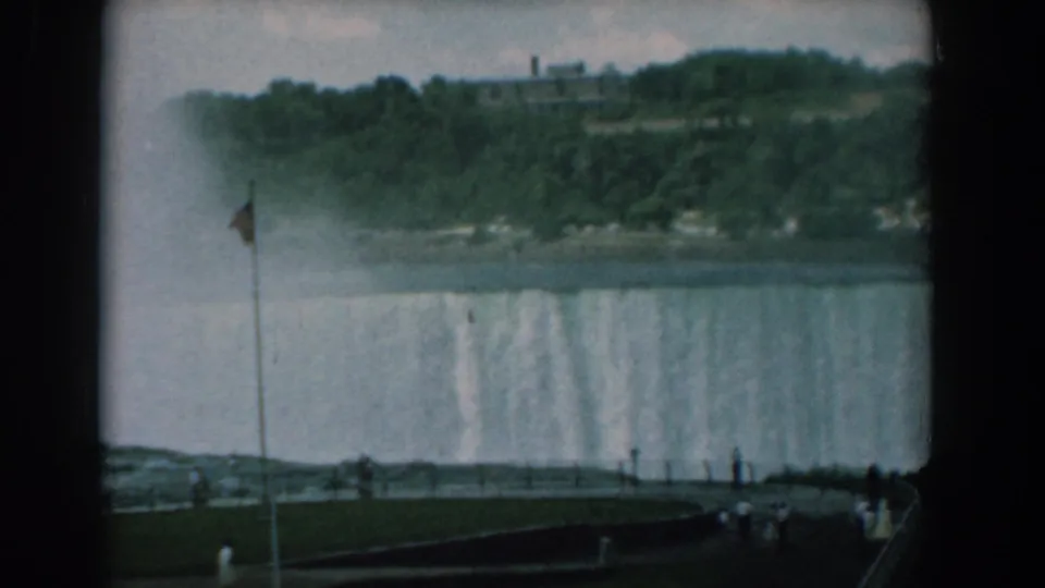 1958 8mm frame of a large waterfall seen from a public overlook, with mist, a flagpole, trees, and tourists in the foreground.