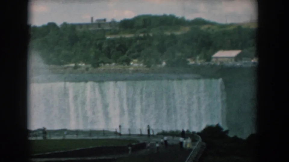 1958 8mm frame showing the broad waterfall rim below a wooded hillside, with a walkway and visitors in the foreground.