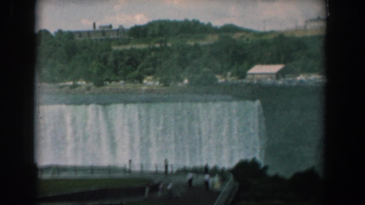 Wide waterfall with tourists along a railing and wooded banks beyond the river.
