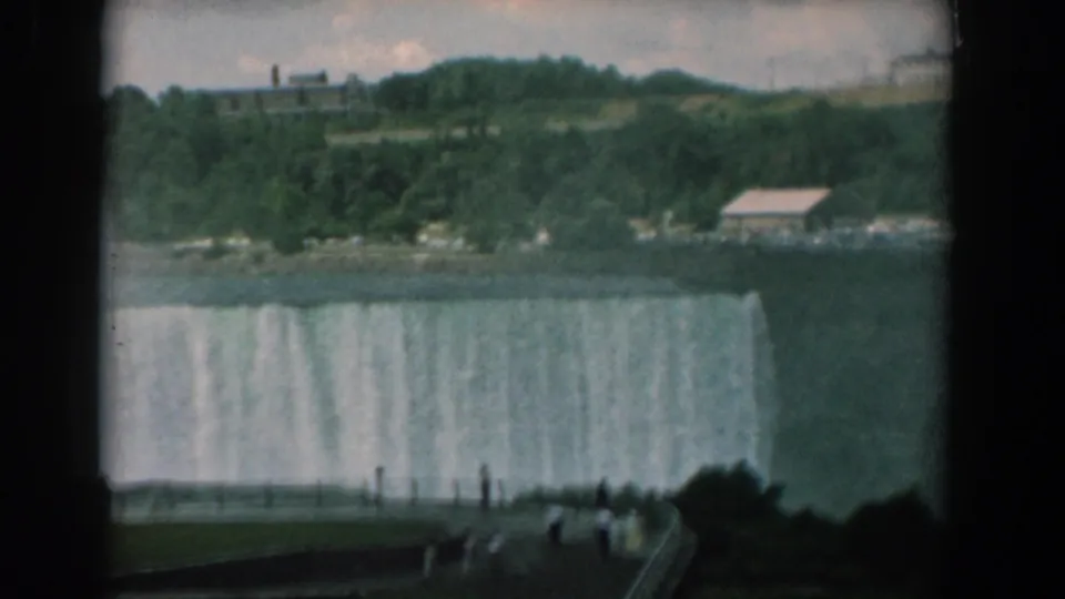 1958 8mm home-movie frame showing tourists at an overlook watching a broad waterfall, with green hills and distant buildings across the water.