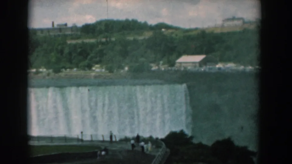 Wide 8mm view of visitors gathered at a railing while a large waterfall drops behind them under an overcast sky.