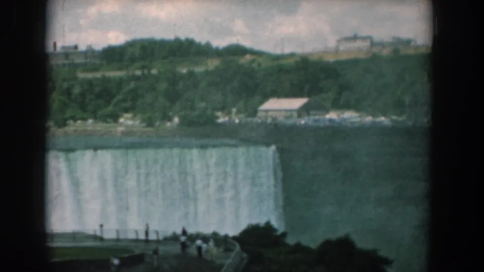Soft 1950s 8mm frame of a waterfall dropping behind an overlook, with visitors in the foreground and wooded slopes beyond.