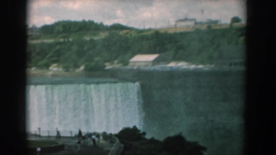 Final 8mm frame of distant tourists viewing a broad waterfall, with a green riverbank, low buildings, and dark home-movie frame borders.