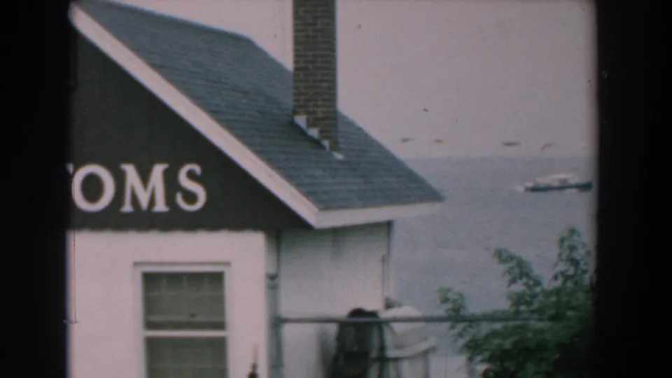 1958 Yorktown home-movie frame showing part of a CUSTOMS sign on a small building near calm water with a distant boat.