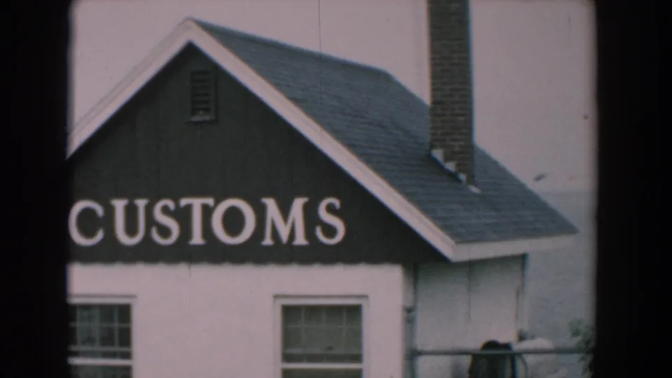 1958 Yorktown home-movie view of the CUSTOMS sign across a small white waterfront building with roof, chimney, windows, and water behind.