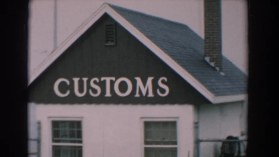 1958 Yorktown frame centering the CUSTOMS sign on the small waterfront building, with chimney, windows, railing, and calm water visible.