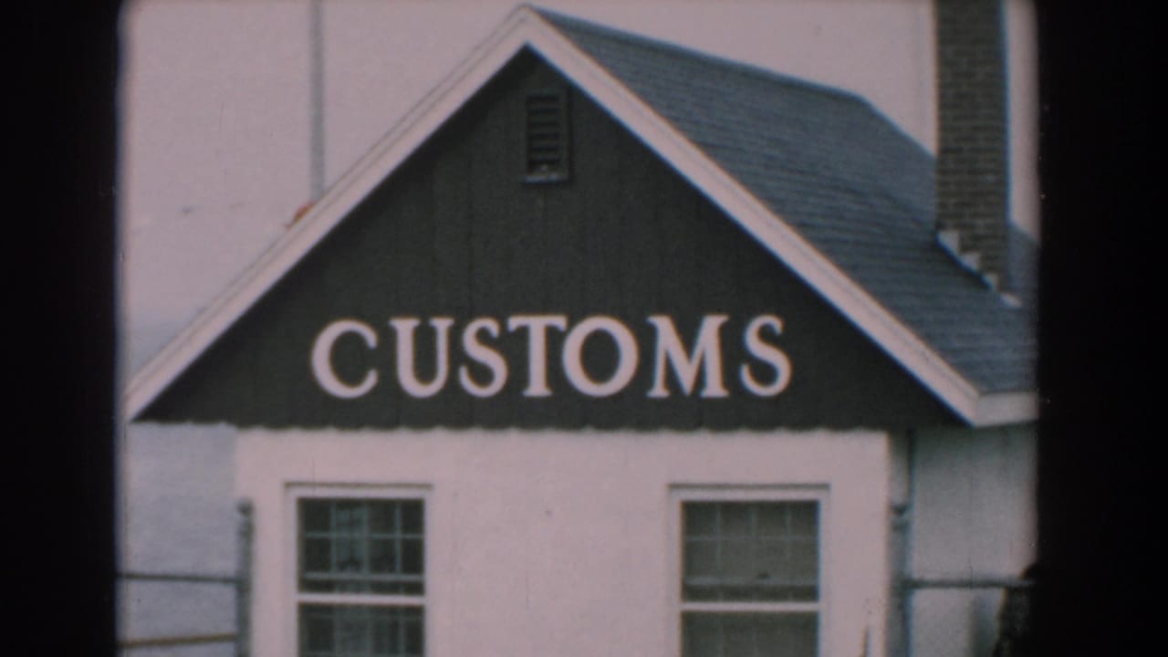 White building exterior with a dark gable marked CUSTOMS, two windows, railing, and water behind it.