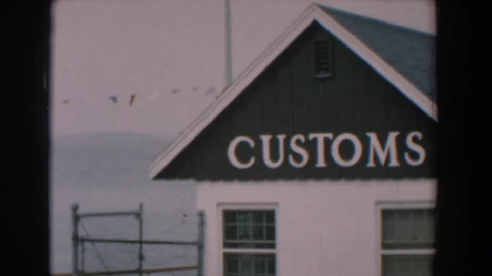 1958 Yorktown home-movie frame cropped on the CUSTOMS sign and white building, with a window, waterfront railing, water, and small pennant flags.