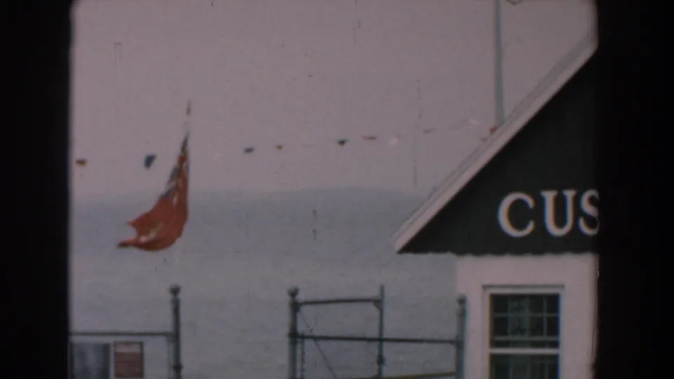 1958 Yorktown, Virginia 8mm frame showing a red flag, chain-link fence, window and cropped CUSTOM lettering on a waterfront building.