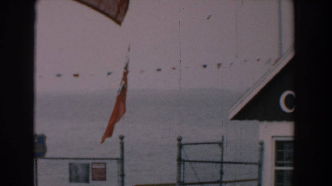 1958 Yorktown, Virginia shoreline view with a red flag, chain-link fence, small signs, pennant string, and gray water in the background.