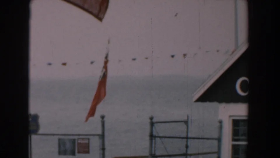 1958 Yorktown, Virginia shoreline view with a red flag, chain-link fence, small signs, pennant string, and gray water in the background.
