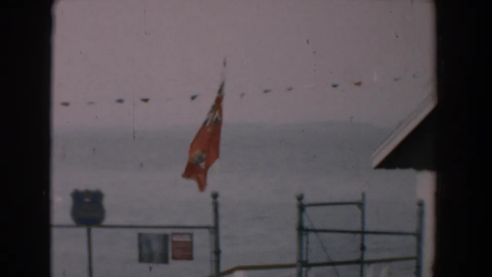 Yorktown shoreline frame from 1958 showing a red flag above fencing, a blue shield-shaped sign, small notice sign, roof edge, and calm water.