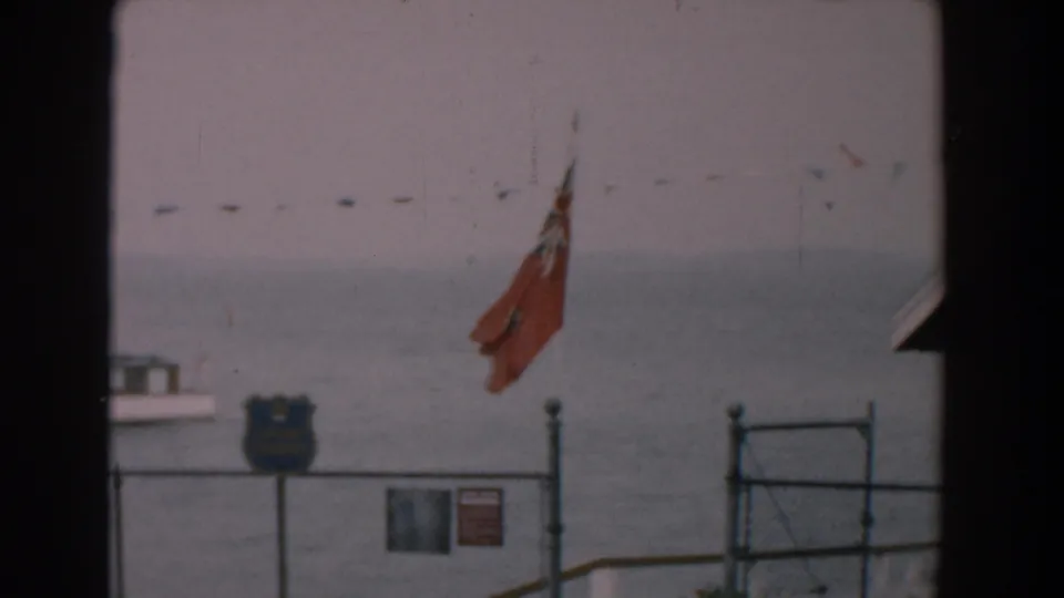 1958 Yorktown waterfront view with the red flag centered, chain-link fence, blue shield-shaped sign, small sign, pennants, and muted water beyond.