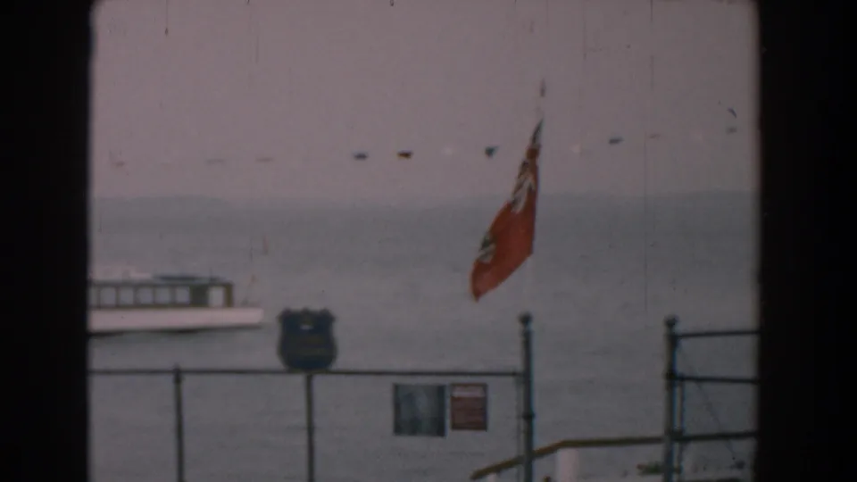 Late-frame 1958 Yorktown shoreline view with the red flag to the right, fencing, blue shield-shaped sign, small sign, roofline, and gray water.
