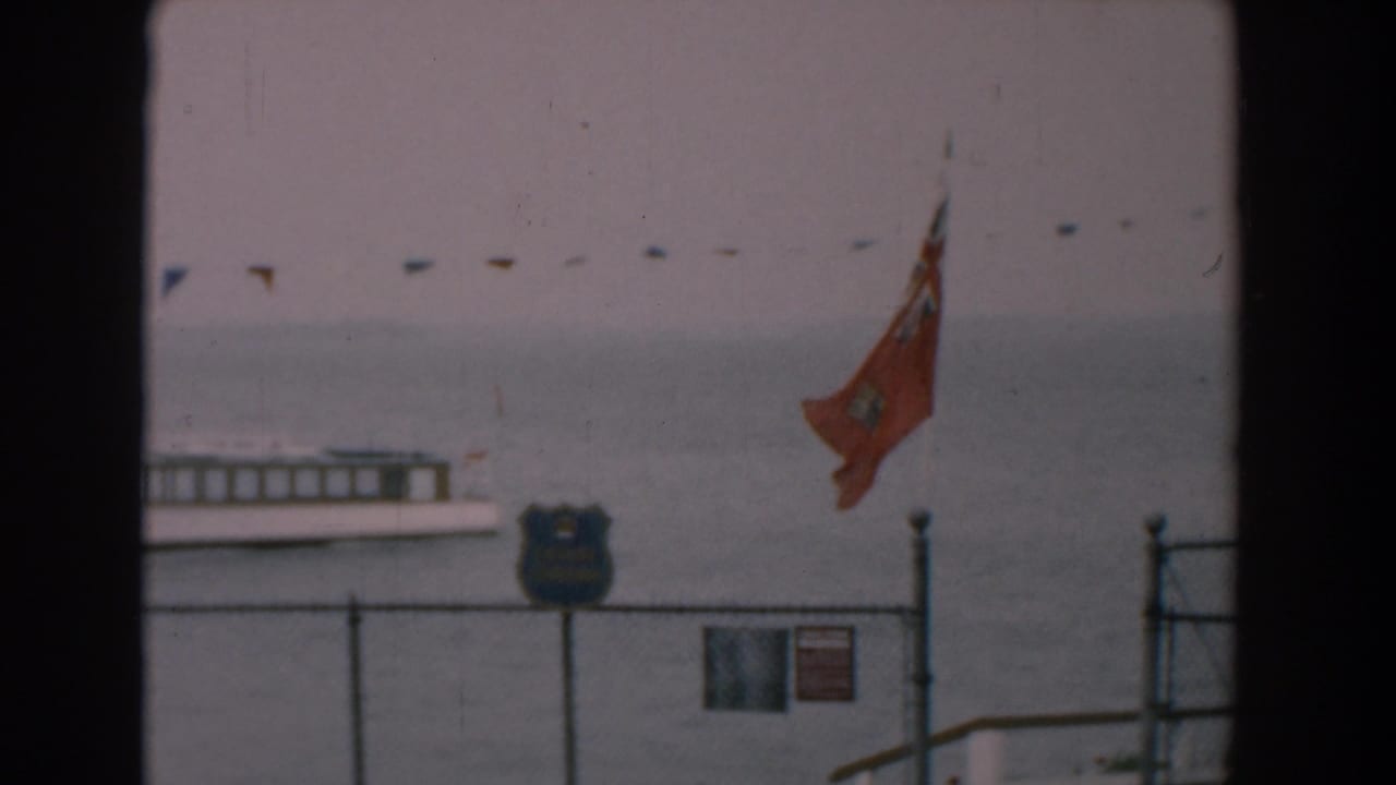 Ferry on calm water behind a fence with a red flag and shoreline signs.