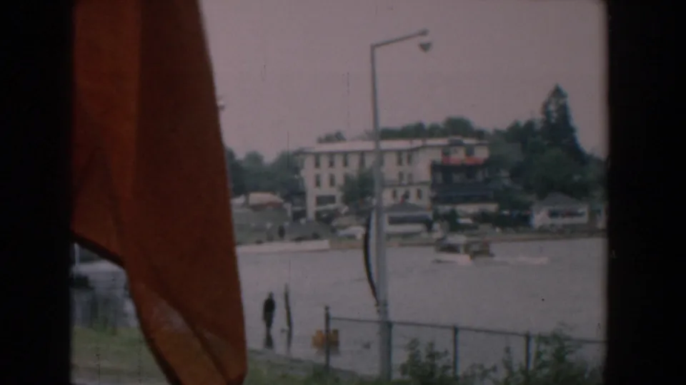 Yorktown waterfront frame with a red flag close to camera, distant buildings, parked cars, shoreline, water, and a small person near the landing.