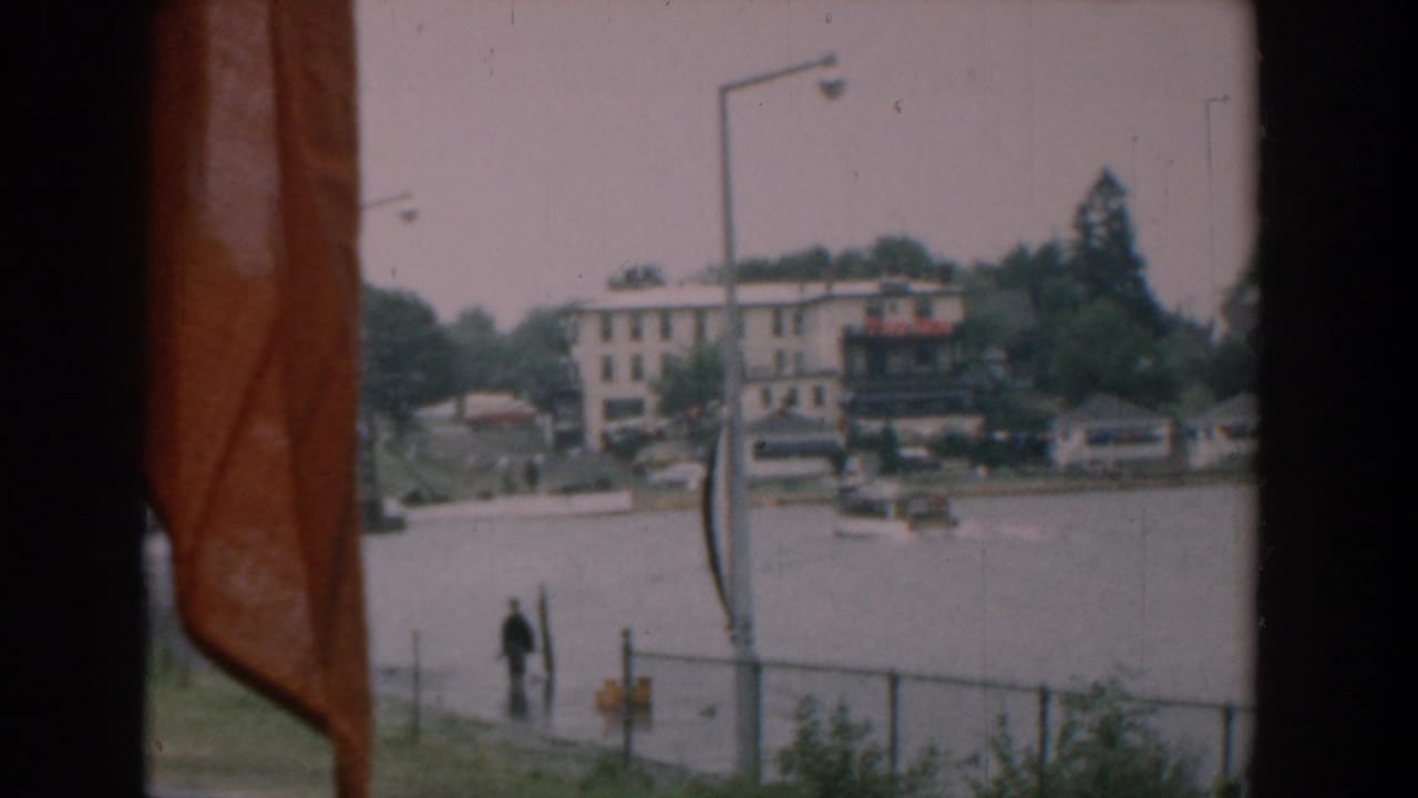 Waterfront seen through dark window edges with orange flag fabric, a pedestrian, boats, fence, and buildings across the river.