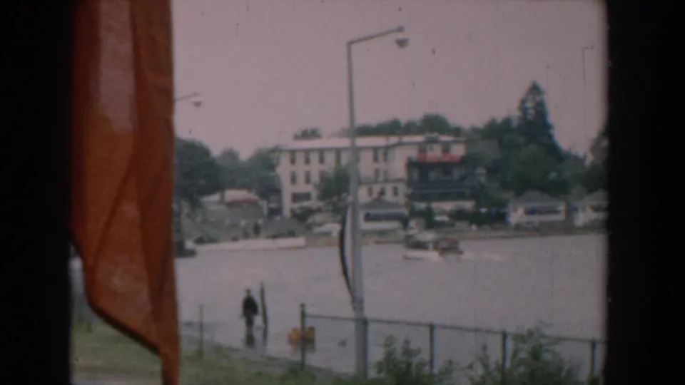 1958 Yorktown waterfront view with orange flag fabric in the foreground, a lone pedestrian near a chain-link fence, boats, streetlights, and buildings across the river.
