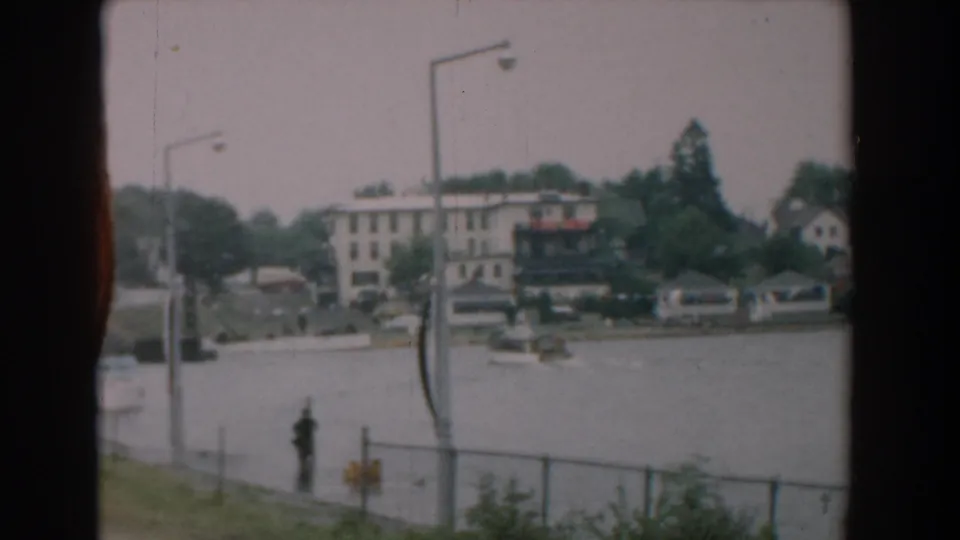 1958 Yorktown riverfront with a lone pedestrian by the fence, small boats near the dock area, tall streetlights, trees, and shoreline buildings.