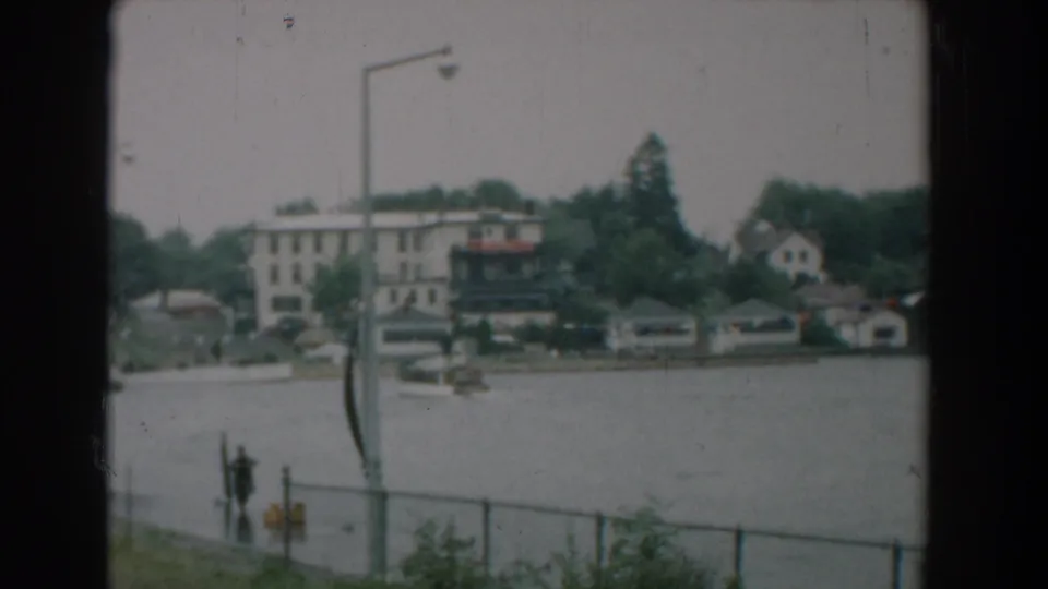 Soft 1958 Yorktown waterfront frame showing a pedestrian near the lower-left fence, river traffic, streetlights, trees, and buildings along the opposite shore.