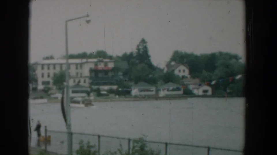 1958 Yorktown waterfront view with a pedestrian near a yellow post, chain-link fence in front, broad river water, boats, trees, and shoreline buildings.