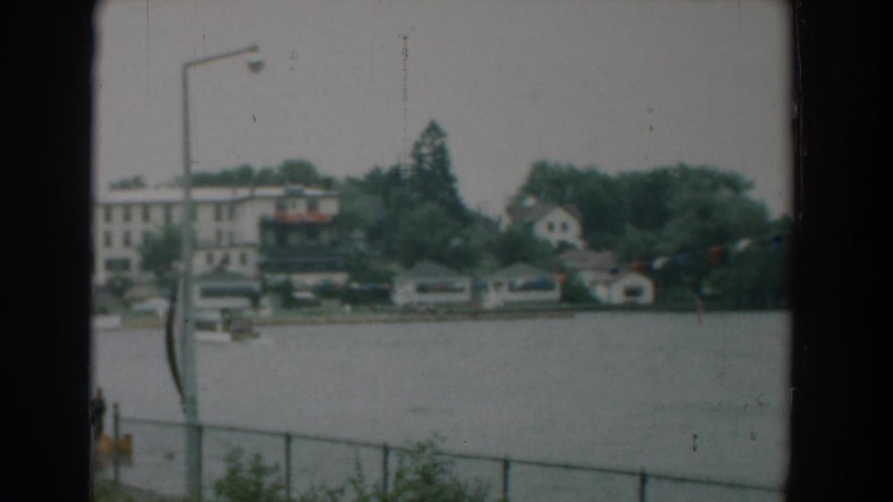 Waterfront view across a lake with houses, trees, boathouses, a railing, and a lamp post in front.