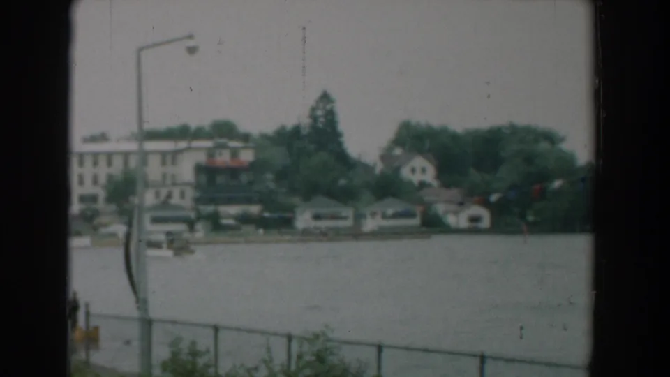 1958 Yorktown, Virginia home-movie frame showing water, a distant residential shore, boathouses, trees, a railing, and a tall lamp post.