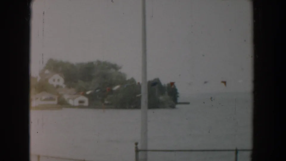 Soft 1958 Yorktown 8mm frame with a rain-muted shoreline, distant houses, trees, gray water, railing, and small colorful flags.