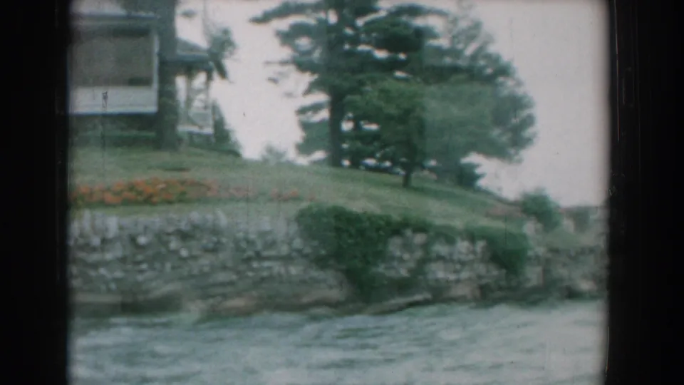 Yorktown riverside view with a grassy slope, stone retaining wall, orange flowers, evergreen trees, water, and a partial house above the bank.
