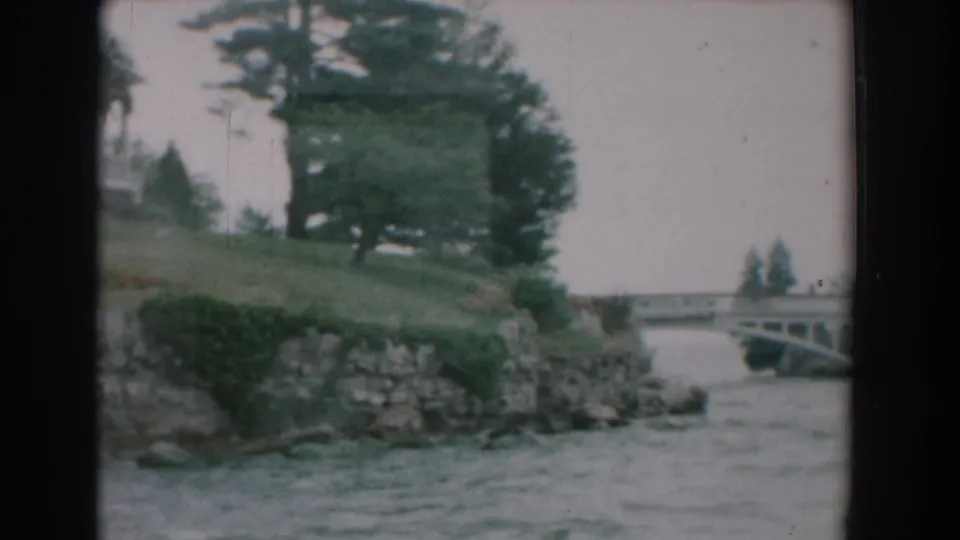 1958 Yorktown river scene with a stone bank, grassy hillside, evergreens, choppy water, and a small bridge in the distance.