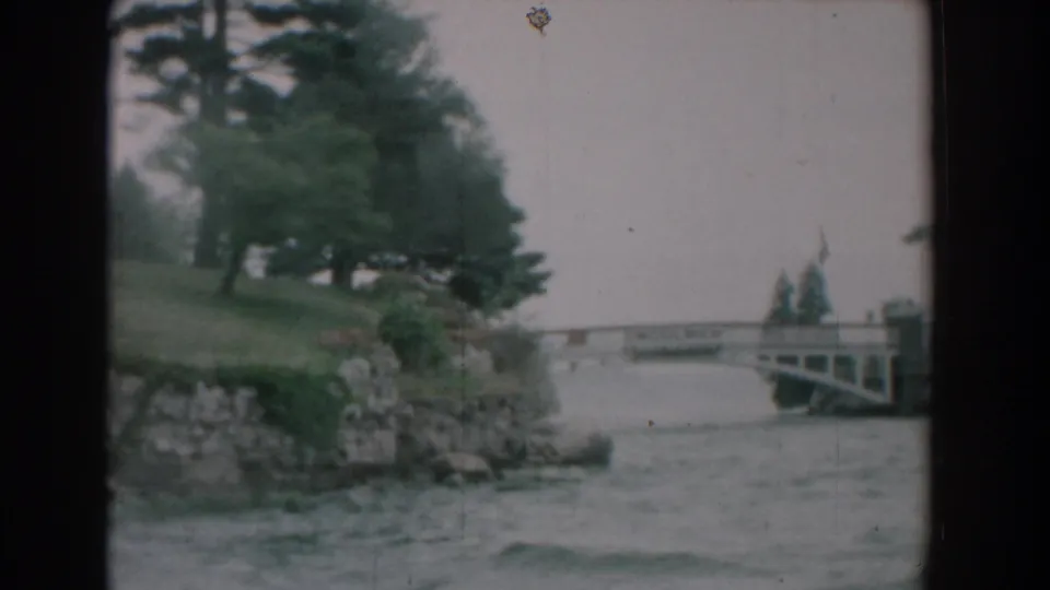 Yorktown waterfront view from 1958 8mm footage, showing evergreens, a stone shoreline, choppy river water, and a small bridge beyond.