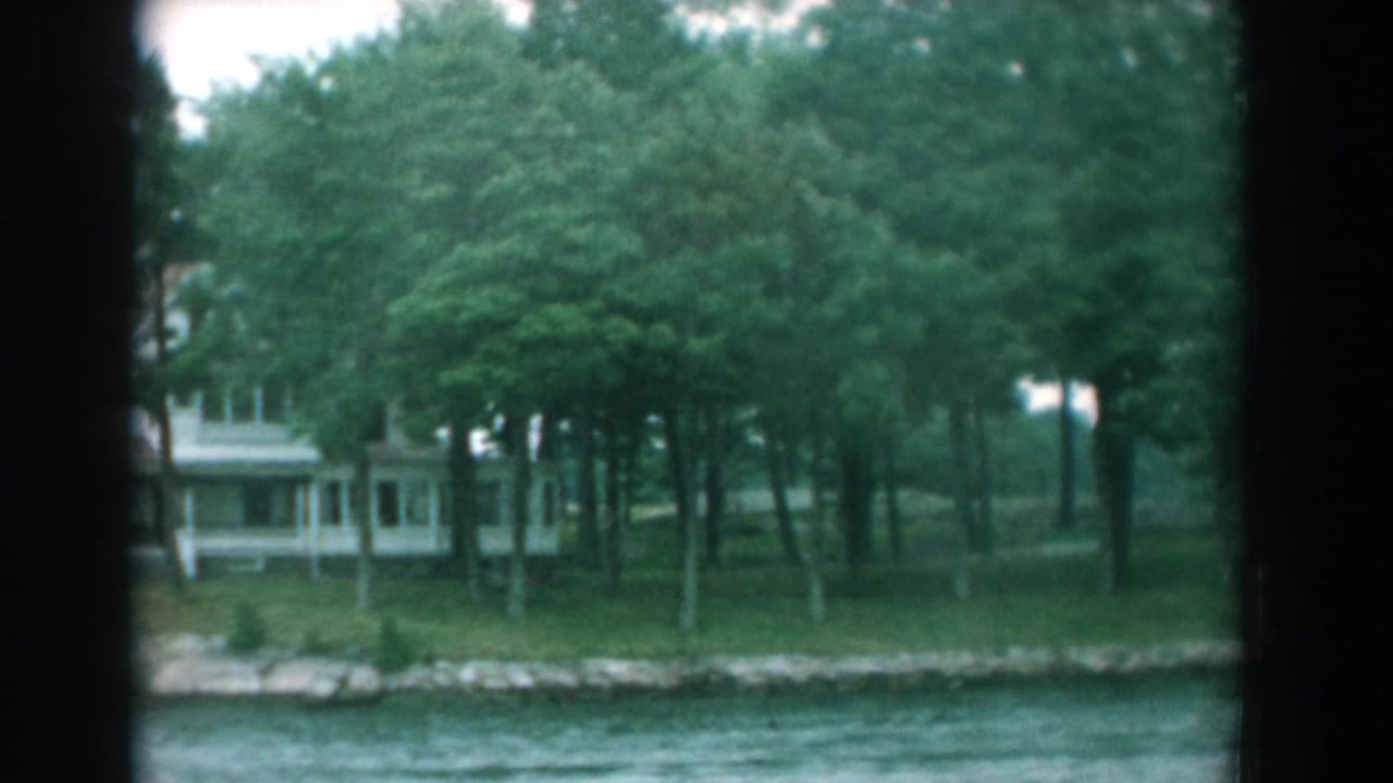Wooded shoreline with rippling water, a pale house, and a low stone wall.