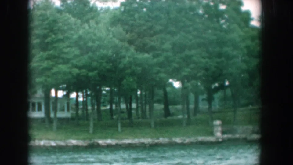 Boat-view frame of a tree-lined Yorktown shore in 1958, with water foreground and a pale building partly hidden by leaves.