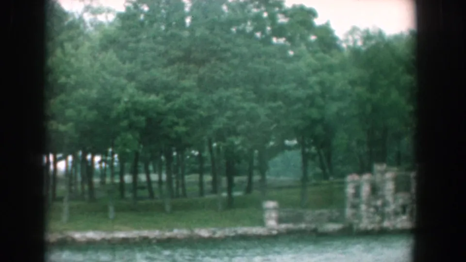 1958 Yorktown shore seen from a moving boat, with dense trees, grassy bank, water, and small stonework near the edge.
