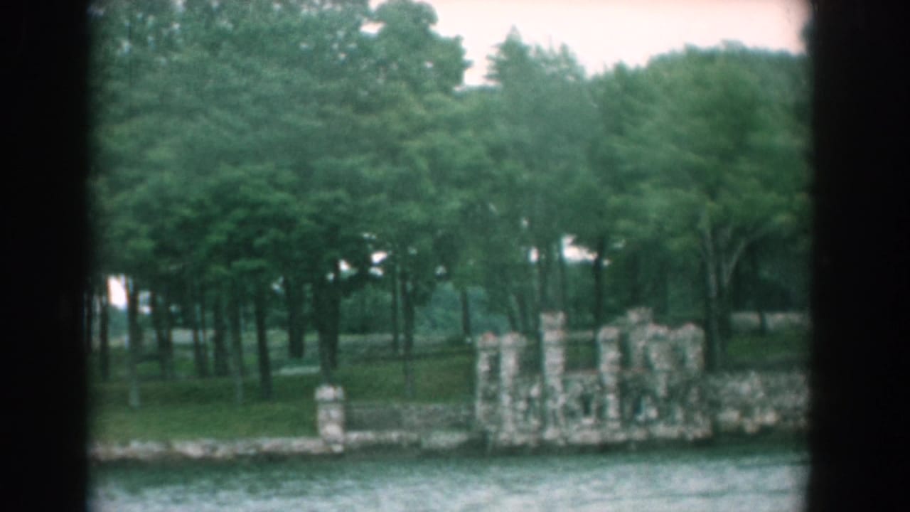 Wooded shoreline with stone wall and ruins seen across moving water.