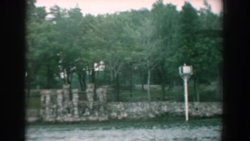 1958 8mm frame from Yorktown showing a stone waterfront wall, wooded background, and a white signpost near the shore.
