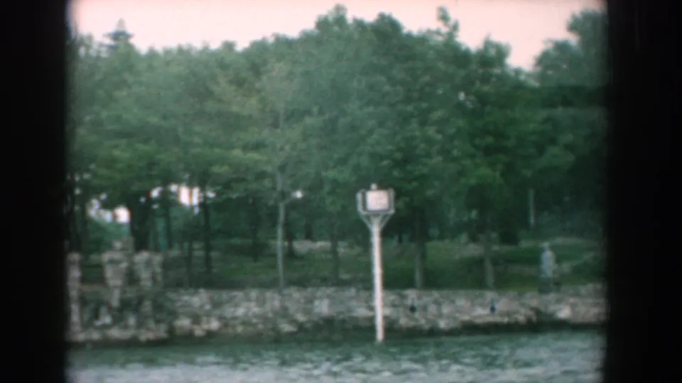 Yorktown-area 1958 home-movie frame with a white shore sign, wooded parkland, stone wall, and rippled water.