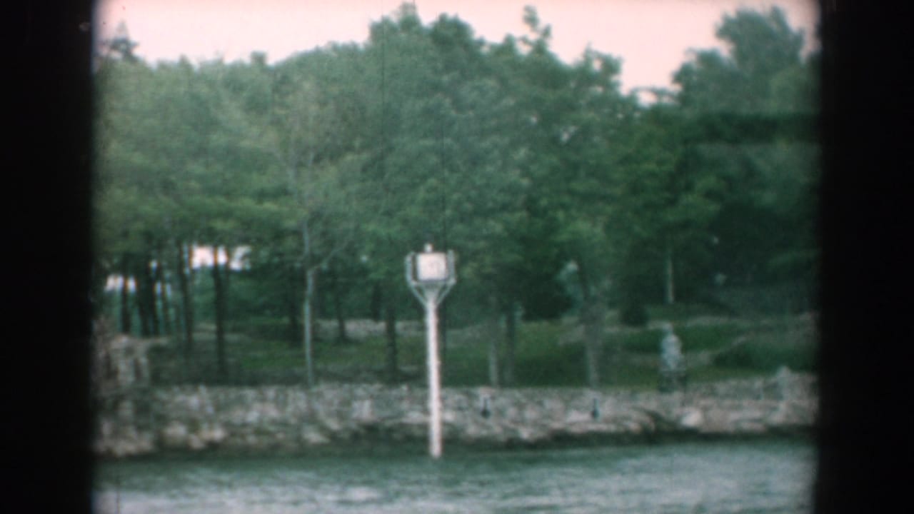 Rocky shoreline with trees, water, and a white post sign seen from a boat.