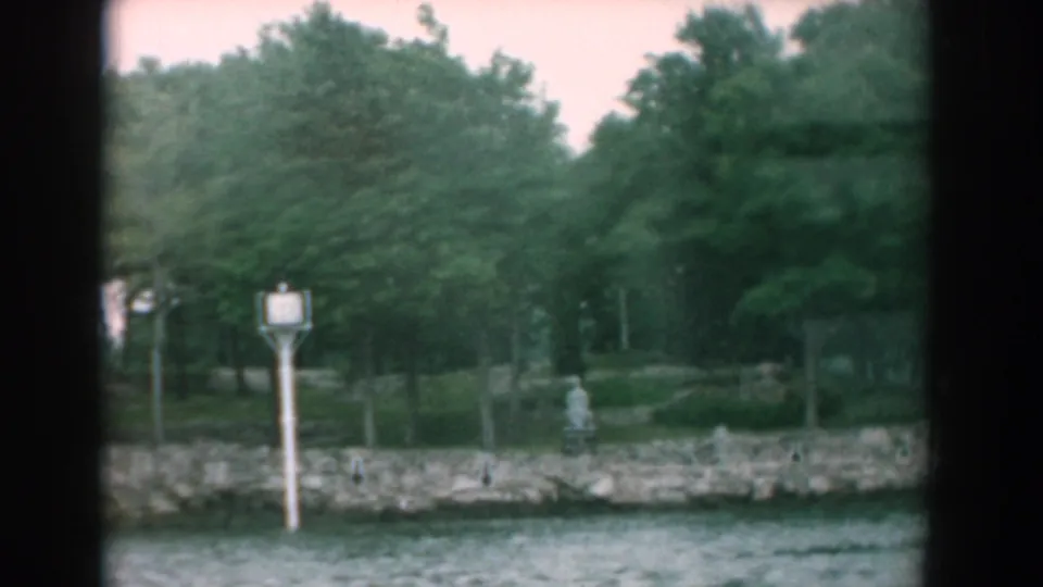 1958 Yorktown, Virginia riverbank view with stone shoreline, dense trees, and a white waterfront signpost filmed from the boat.