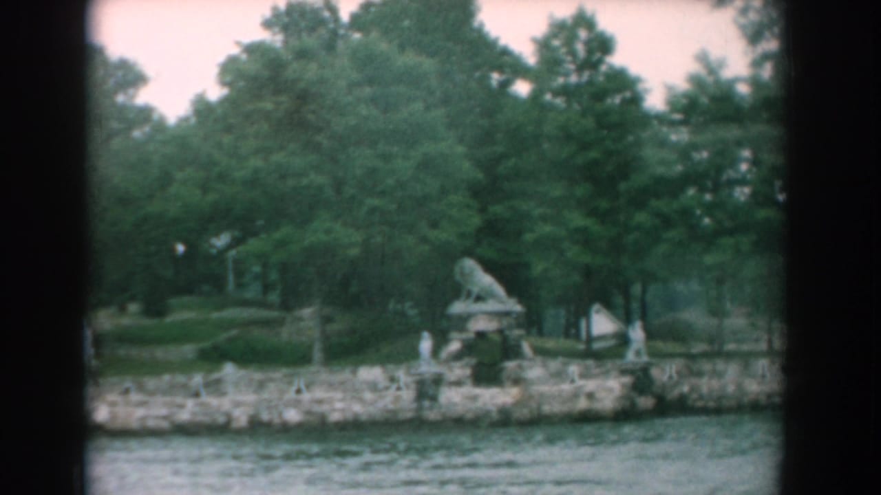 Waterfront with trees, stone shoreline, small statues, and a lion sculpture seen from across the water.