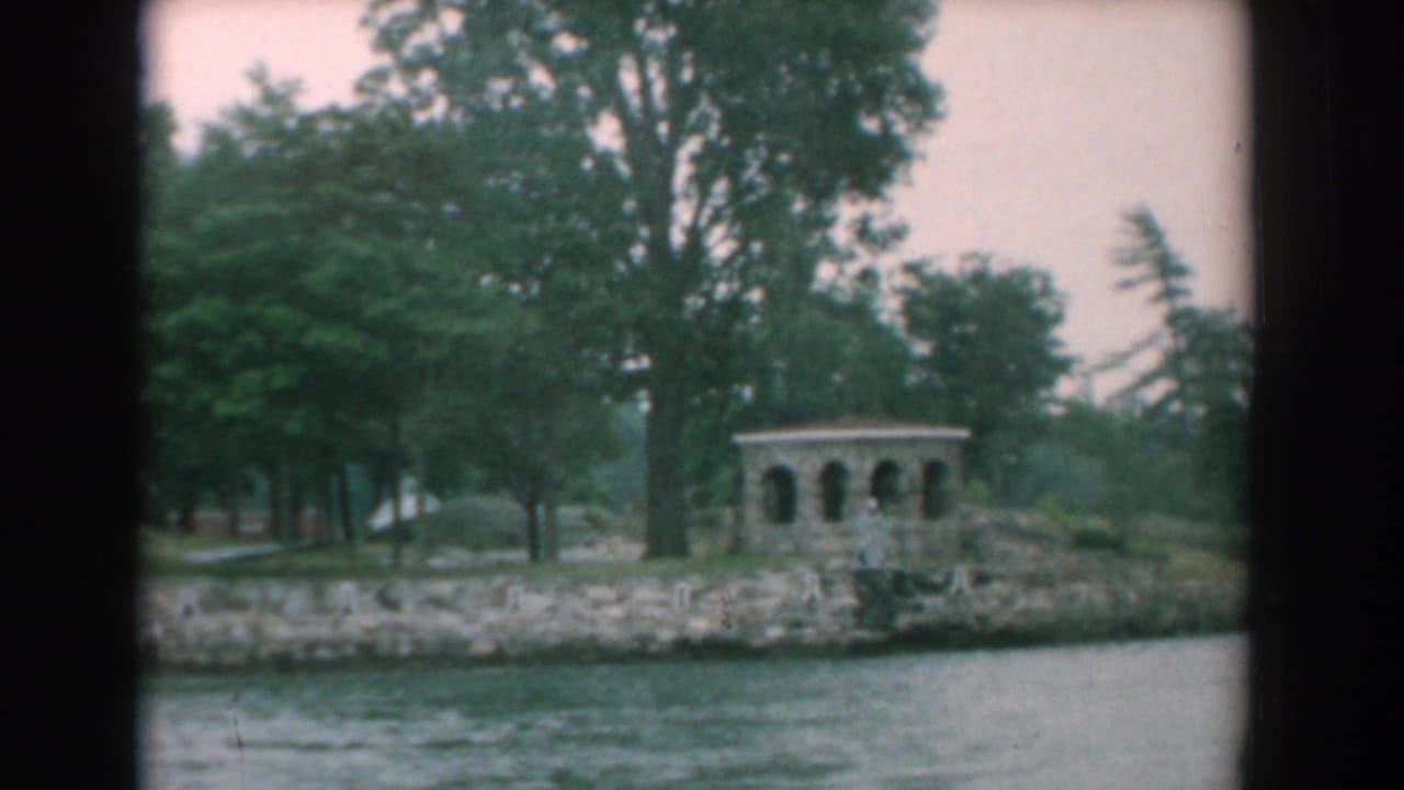 River shoreline with trees, rocky bank, and a small arched gazebo seen from the water.