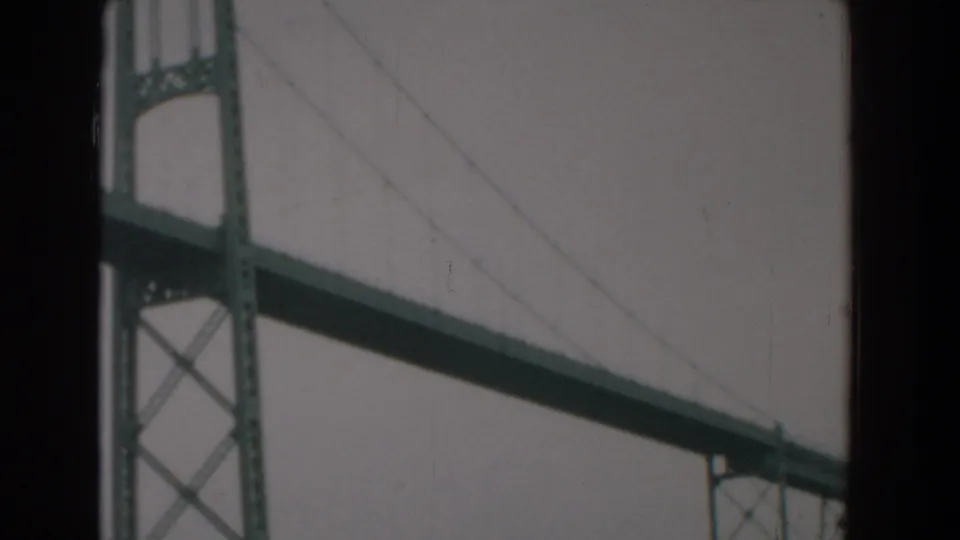 Yorktown, Virginia 1958 home-movie frame with a long suspension bridge deck, tower section, and faint cables against gray sky.
