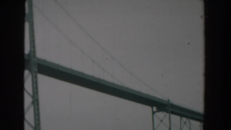 Faded 8mm view of the Yorktown bridge structure, with partial tower framing, diagonal cables, and a long deck under cloud cover.