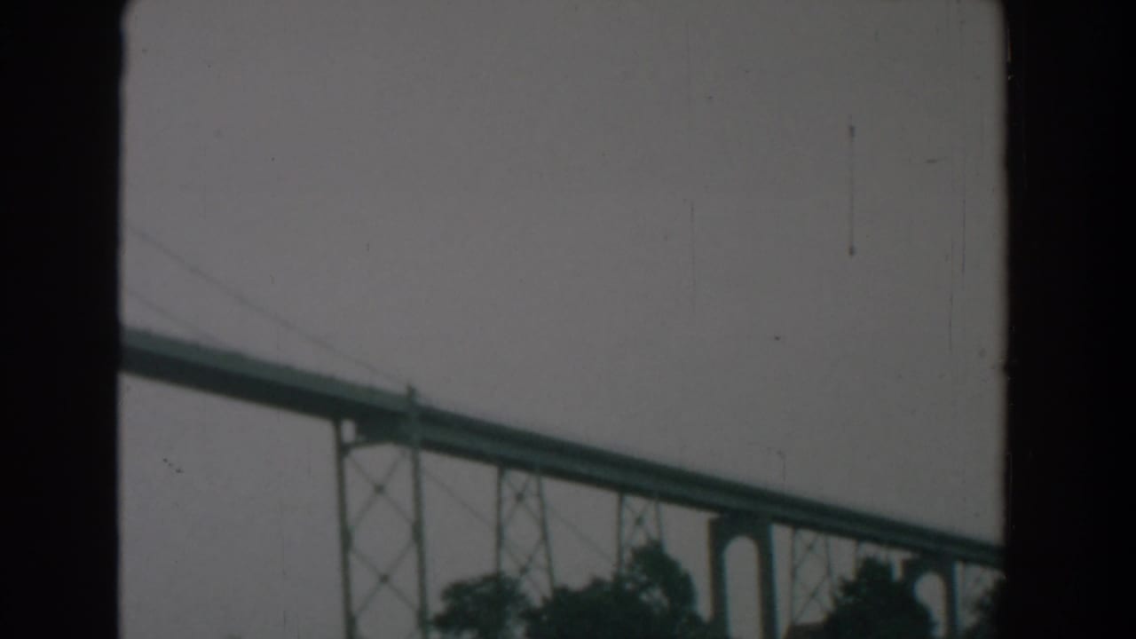 Green bridge span above treetops under a gray sky, framed by dark film borders.