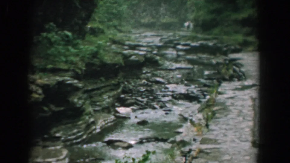 1958 Yorktown, Virginia 8mm frame showing a shallow stream flowing through layered rocks, with trees and a faint distant figure beyond the channel.